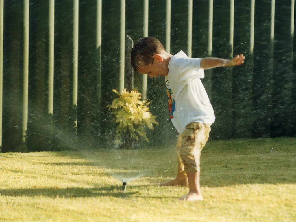 A smart irrigation controller mounted on a wall with a sprinkler watering a lawn in the background